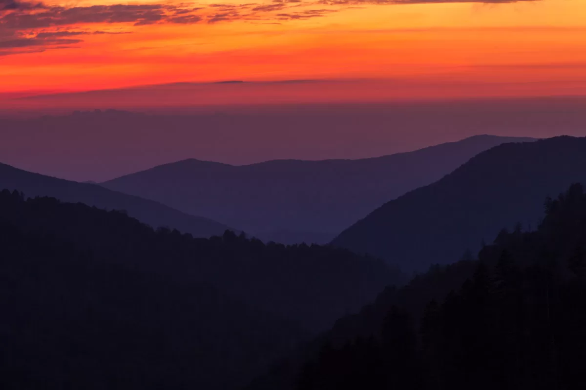 This is a photo of a stunning sunset over a mountain range. The sky is ablaze with vibrant colors of orange, pink, and purple, creating a breathtaking backdrop. The mountains below are silhouetted against the colorful sky, their dark shapes contrasting beautifully with the bright colors above. The overall atmosphere of the image is one of peace, tranquility, and awe-inspiring beauty.